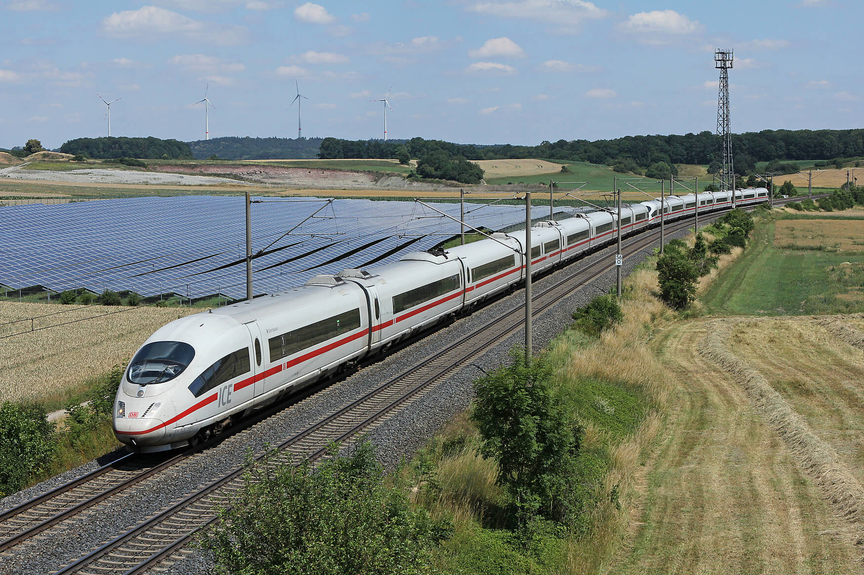 ICE 4 fährt an Solarpark vorbei, im Hintergrund Wald und Windräder